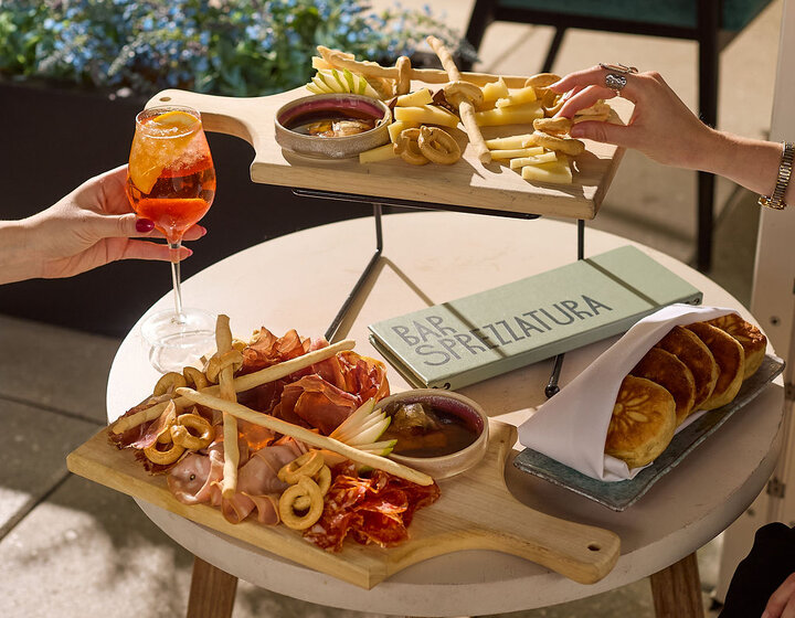a hand holding a sprtiz and another hand taking a snack from a charcuteries plate at Bar Sprezzatura with other snack plates  untouched
